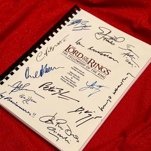 May include: A white book with the title "The Lord of the Rings: The Fellowship of the Ring" and several autographs in blue ink. The book is lying on a red surface.
