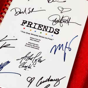 May include: A white script with the title "FRIENDS" and the episode title "THE ONE WHERE ROSS IS FINE". The script is covered in signatures in black and blue ink. The background is a red, glittery surface.