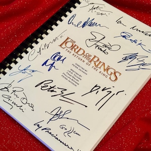 Peut inclure: Un livre blanc avec le titre "Le Seigneur des Anneaux : Le Retour du Roi" et plusieurs signatures à l'encre noire. Les signatures incluent "Peter Jackson", "Fran Walsh", "Philippa Boyens", "Gollum", "Gimli", et d'autres.