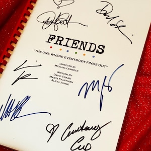 Puede incluir: Un guion blanco con el título "FRIENDS" y el título del episodio "THE ONE WHERE EVERYBODY FINDS OUT". El guion está firmado con tinta negra y azul. El guion tiene una encuadernación en espiral roja. El fondo es rojo.