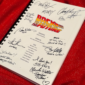 Puede incluir: Un guion blanco con el título "Regreso al futuro" en rojo y naranja. El guion está firmado por el reparto y el equipo, incluidos Michael J. Fox, Christopher Lloyd, Lea Thompson y Crispin Glover. Las firmas están en tinta negra.