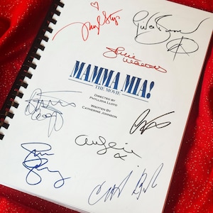 Könnte beinhalten: Ein weißer spiralgebundener Notizbuchumschlag mit dem blauen Text Mamma Mia! The Movie, Directed by Phyllidia Lloyd, Written by Catherine Johnson, umgeben von mehreren handschriftlichen Unterschriften in schwarzer, blauer und roter Tinte, inklusive eines kleinen Herzens.