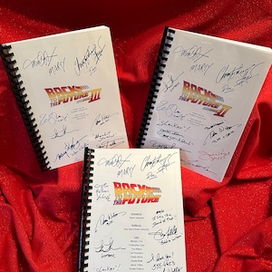 May include: Three white spiral bound screenplays for the movie Back to the Future. The covers are signed by the cast and crew, including Michael J. Fox, Christopher Lloyd, Lea Thompson, and others. The screenplays are on a red surface.