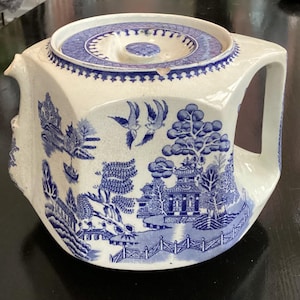 Könnte beinhalten: Eine quadratische Vintage-Teekanne mit blau-weißem Willow-Muster. Die Teekanne hat einen Deckel und einen Griff. Das Muster umfasst Szenen von Pagoden, Brücken, Bäumen und Vögeln. Die Teekanne ist aus Keramik.