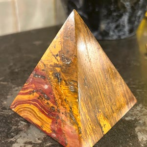 May include: A pyramid-shaped crystal with red, orange, and yellow stripes. The crystal is polished and has a smooth surface.