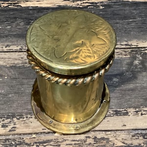 May include: A vintage brass container with a lid featuring an embossed floral design. The cylindrical body is accented with a rope-like trim and sits on a circular base. The container is made of brass.