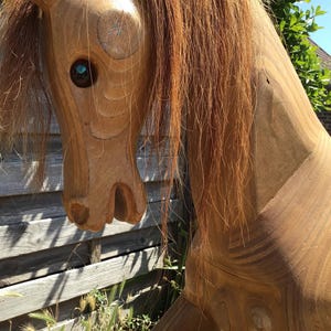 May include: A detailed wooden rocking horse head, showing intricate carving and a light brown mane. The horse has a dark eye and is set against a backdrop of a wooden fence and greenery. The wood grain is visible.
