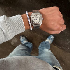 Könnte beinhalten: Eine silberfarbene, quadratische Armbanduhr mit weißem Zifferblatt, schwarzen römischen Ziffern und blauen Zeigern, kombiniert mit einem Perlenarmband mit blau-weißem Blumenmuster. Die Uhr hat ein Metallgliederarmband und eine sichtbare Krone.