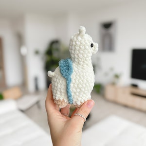 May include: A white crocheted llama toy with a blue and brown saddle. The llama has black eyes and a white fluffy body.