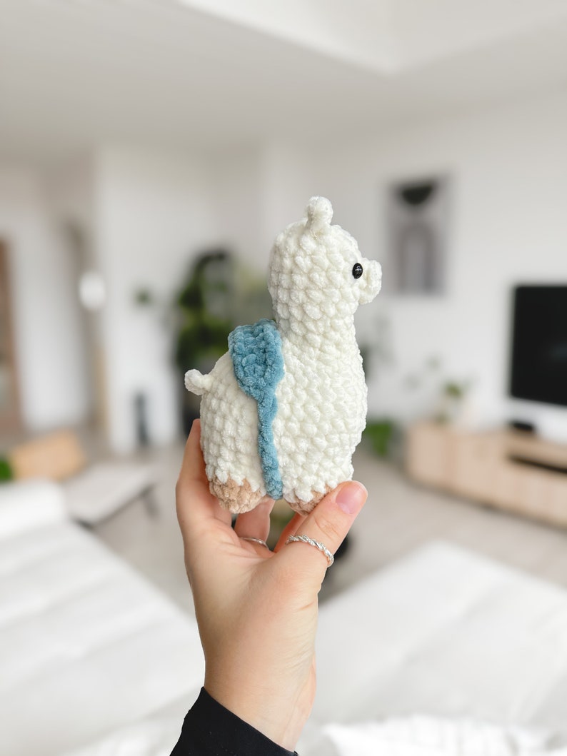 May include: A white crocheted llama toy with a blue and brown saddle. The llama has black eyes and a white fluffy body.