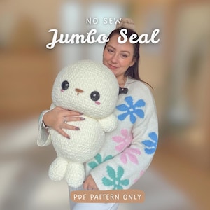 Könnte beinhalten: Ein großes, cremefarbenes gehäkeltes Seehund-Plüschtier mit schwarzen Augen und einer braunen Nase wird von einer Person gehalten. Der Text "NO SEW Jumbo Seal" und "PDF PATTERN ONLY" sind auf dem Bild zu sehen. Die Person trägt einen weißen Pullover mit floralen Mustern.