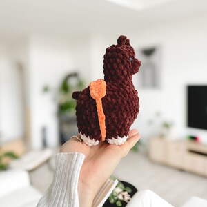 May include: A brown crocheted llama toy with orange accents. The llama is standing on its four legs and has a fluffy texture.