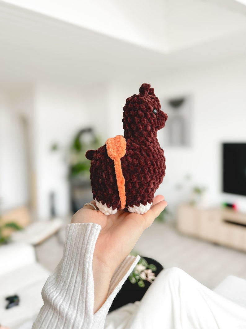 May include: A brown crocheted llama toy with orange accents. The llama is standing on its four legs and has a fluffy texture.