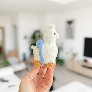 May include: A white fluffy alpaca plush toy with a blue and white striped scarf. The toy has brown feet and is being held by a hand.