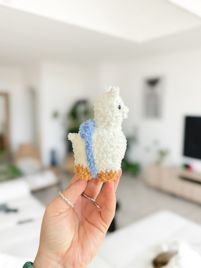 May include: A white fluffy alpaca plush toy with a blue and white striped scarf. The toy has brown feet and is being held by a hand.