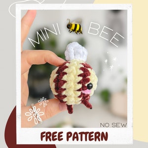 May include: A crocheted bee toy with a white pom-pom on its head. The bee is brown and yellow and has a black face with a pink tongue. The bee is being held in a hand and the text "MINI BEE FREE PATTERN NO SEW" is visible below the bee.