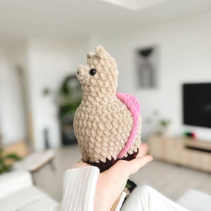 May include: A beige crocheted llama plush toy with pink and brown accents. The llama is being held by a person's hand.