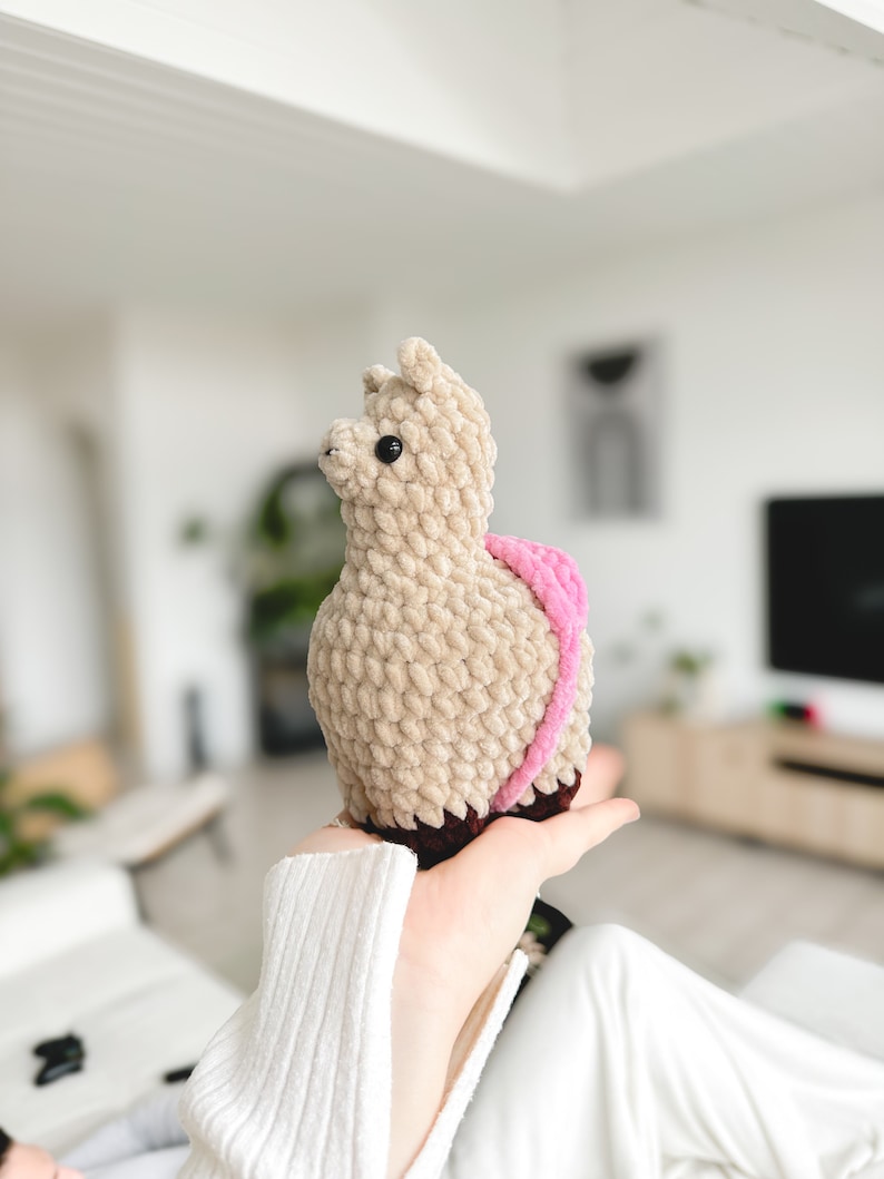 May include: A beige crocheted llama plush toy with pink and brown accents. The llama is being held by a person's hand.