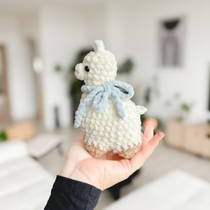 May include: A white crocheted llama with a light blue scarf. The llama is standing on a person's hand.