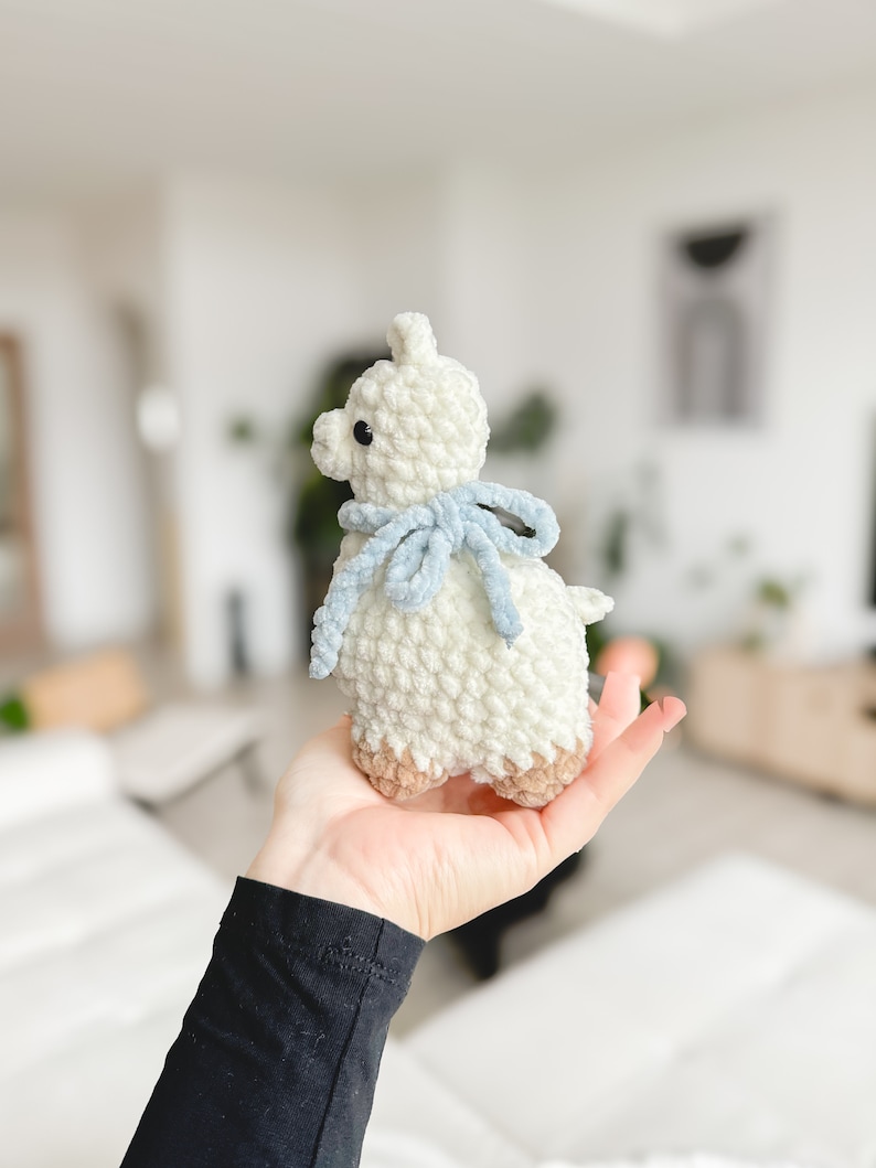 May include: A white crocheted llama with a light blue scarf. The llama is standing on a person's hand.