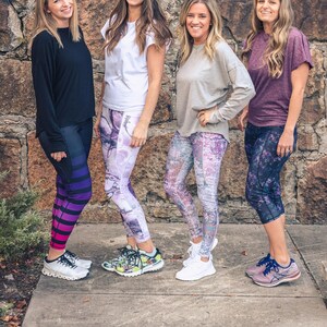 May include: Four women wearing different patterned leggings and tops stand in front of a stone wall. They are all smiling and looking at the camera. The leggings are all different colors and patterns, and the tops are all different colors and styles.