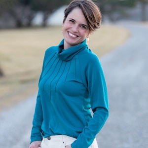 Peut inclure: Une femme portant un pull col roulé bleu turquoise avec un pantalon blanc. Elle se tient sur un chemin de gravier avec des arbres en arrière-plan.