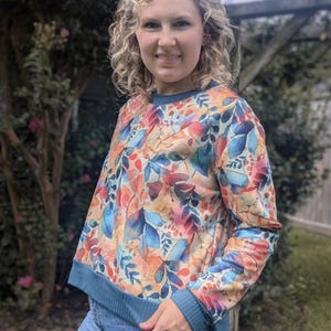 May include: A long-sleeved, floral print sweatshirt with a teal blue ribbed neckline and cuffs. The sweatshirt has a watercolour-like design with shades of blue, orange, and pink.