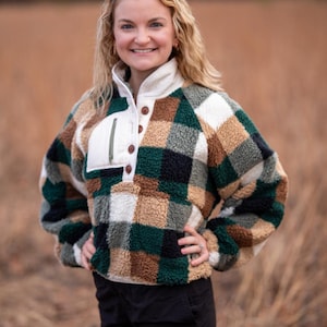 Peut inclure: Une femme portant un pull en polaire à carreaux vert, noir, blanc et marron avec une poche blanche et des boutons marrons. Elle porte un short noir et se tient dans un champ d'herbe haute.