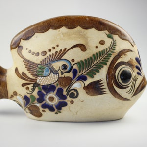 May include: A ceramic fish figurine with a brown, white, and blue painted design. The fish has a bird with green, blue, and white wings painted on its side.