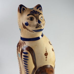 May include: A ceramic cat figurine with brown, blue, and white painted details. The cat is sitting upright with its tail curled around its body.