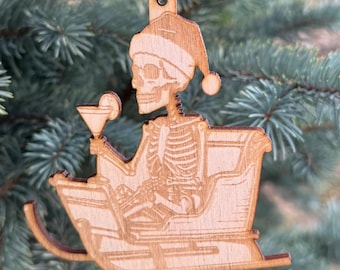 Puede incluir: Adorno navideño de madera que representa un esqueleto con un gorro de Papá Noel, sosteniendo un cóctel en un trineo. El adorno de madera marrón claro cuelga de una cuerda roja y blanca. El adorno está en un pino azul-verdoso.