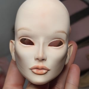 May include: A pale, blank doll head with detailed facial features. The doll head has open eye sockets, a defined nose, and full lips. The skin tone is a light, neutral color with subtle shading. The doll head is held in a hand.