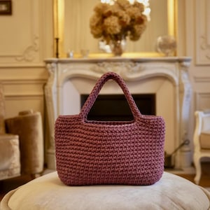 Peut inclure: Un sac fourre-tout en crochet de couleur mauve, de forme rectangulaire, avec une poignée tressée. Le sac est posé sur un pouf rond et rembourré. L'arrière-plan comprend une cheminée, un miroir et un lustre.