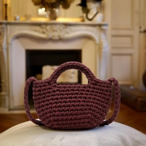 Può includere: Una borsa a mano all'uncinetto bordeaux con base arrotondata e due manici. La borsa è realizzata con un filato spesso e strutturato e poggia su un cuscino di colore chiaro. Lo sfondo comprende un camino ed elementi decorativi.