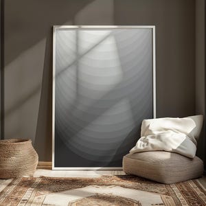 Puede incluir: Una impresión enmarcada en blanco con un diseño abstracto gris y blanco de círculos concéntricos. La impresión está colgada en una pared marrón sobre un cojín de suelo beige con una almohada blanca. Una cesta tejida está en el suelo a la izquierda del cojín.