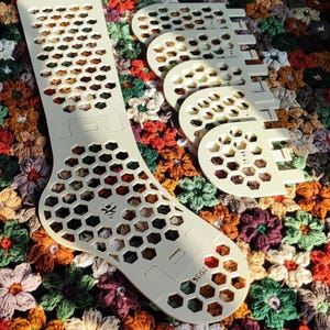 Könnte beinhalten: Ein Set aus hellen, flachen, lasergeschnittenen Sockenblockern aus Holz mit einem sich wiederholenden hexagonalen Wabenmuster. Das Hauptteil ist sockenförmig mit der Aufschrift 35/36, ergänzt durch vier kleinere, abgerundete, halbkreisförmige Einsätze.