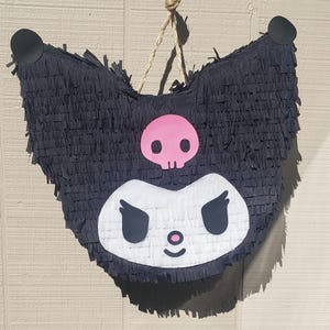 May include: A black piñata in the shape of a cartoon character's head, with a white face, black eyes, and a pink nose. A pink skull is above the face. The piñata is made of fringed paper and has two black circular ears. A rope is attached for hanging.