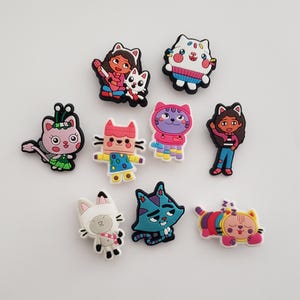May include: A collection of colorful cartoon cat-themed shoe charms. The charms feature various cat characters in different outfits and poses, including a sleeping cat, a cat with a bow, and a cat wearing a sweater. The charms are made of a rubbery material.