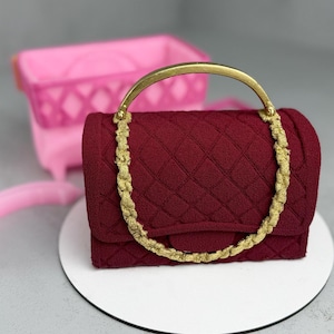May include: A burgundy quilted handbag with a gold-colored handle and chain strap. The bag is displayed on a white circular surface, with a pink basket in the background.