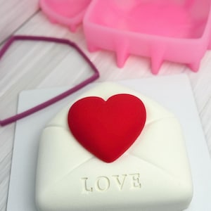 以下が含まれることがあります： 赤いハートが上に乗った、白い封筒型のケーキ。前面には「LOVE」の文字がエンボス加工されています。背景にはピンクのハート型の型と紫色の幾何学的な形があります。ケーキは白い表面にあります。