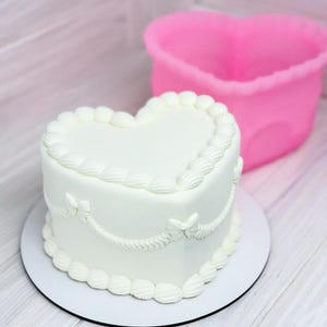 Puede incluir: Un pastel blanco en forma de corazón con glaseado decorativo, sobre un plato blanco. Hay un recipiente rosa en forma de corazón al lado. El fondo es de madera clara.
