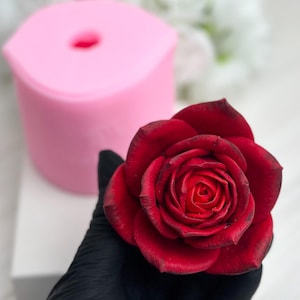 Puede incluir: Una rosa roja vibrante con bordes oscuros se sostiene en una mano enguantada, junto a un molde de silicona rosa. La rosa tiene pétalos detallados y una apariencia realista. El molde es cilíndrico con un agujero en el centro, lo que sugiere un proyecto de manualidades o bricolaje.