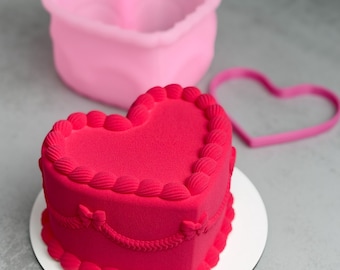 Op de afbeelding: Een hartvormige rode cake met decoratieve frosting op een wit bord. Een roze hartvormige mal en een roze hartvormige koekjesvorm staan op de achtergrond. De cake heeft een fluweelzachte textuur.