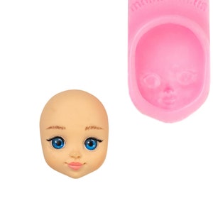 Può includere: Una testa di bambola con occhi blu e uno stampo in silicone rosa di un viso di bambola. La testa della bambola ha sopracciglia, ciglia e labbra dipinte. Lo stampo rosa ha il testo "mold.com.ua" stampato.