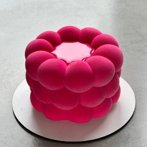 Puede incluir: Un pastel de postre rosa vibrante con un diseño esférico único. El pastel se asienta sobre una base circular blanca. La parte superior del pastel tiene un glaseado rosa brillante. El exterior del pastel es de un rosa profundo y saturado.