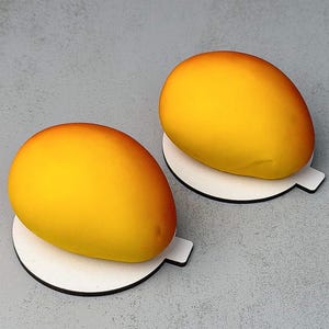 May include: Two smooth, oval-shaped mango-colored desserts with a gradient from bright yellow to orange. Each piece is resting on a small, circular white cardboard base with a protruding tab, presenting a realistic fruit-like appearance.