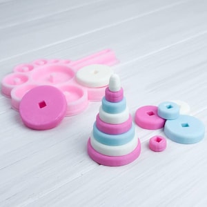 以下が含まれることがあります： ピンクとブルーのプラスチック製の積み重ねおもちゃ。おもちゃは円錐形で、互いに積み重ねることができる複数のリングがあります。おもちゃは白い木製の表面の上に置かれています。