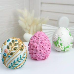 May include: Three decorative Easter eggs. One is light blue with gold and teal leaf designs and small pink and blue flowers. Another is covered in pink flowers. The third is white with green leaves and white flowers.