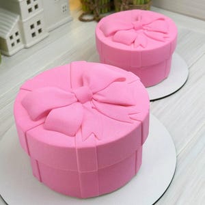 Può includere: Due torte rotonde a forma di scatole regalo, ricoperte di glassa rosa liscia. Ogni torta presenta un fiocco rosa scolpito sulla parte superiore e bande rosa verticali sui lati, che ricordano un nastro, poggiate su basi circolari bianche.