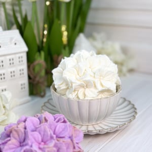May include: A still-life composition featuring a white ceramic bowl filled with white flowers, resting on a matching saucer. A purple hydrangea and white tulips are in the background, with a small white house. The scene is set on a white wooden surface.